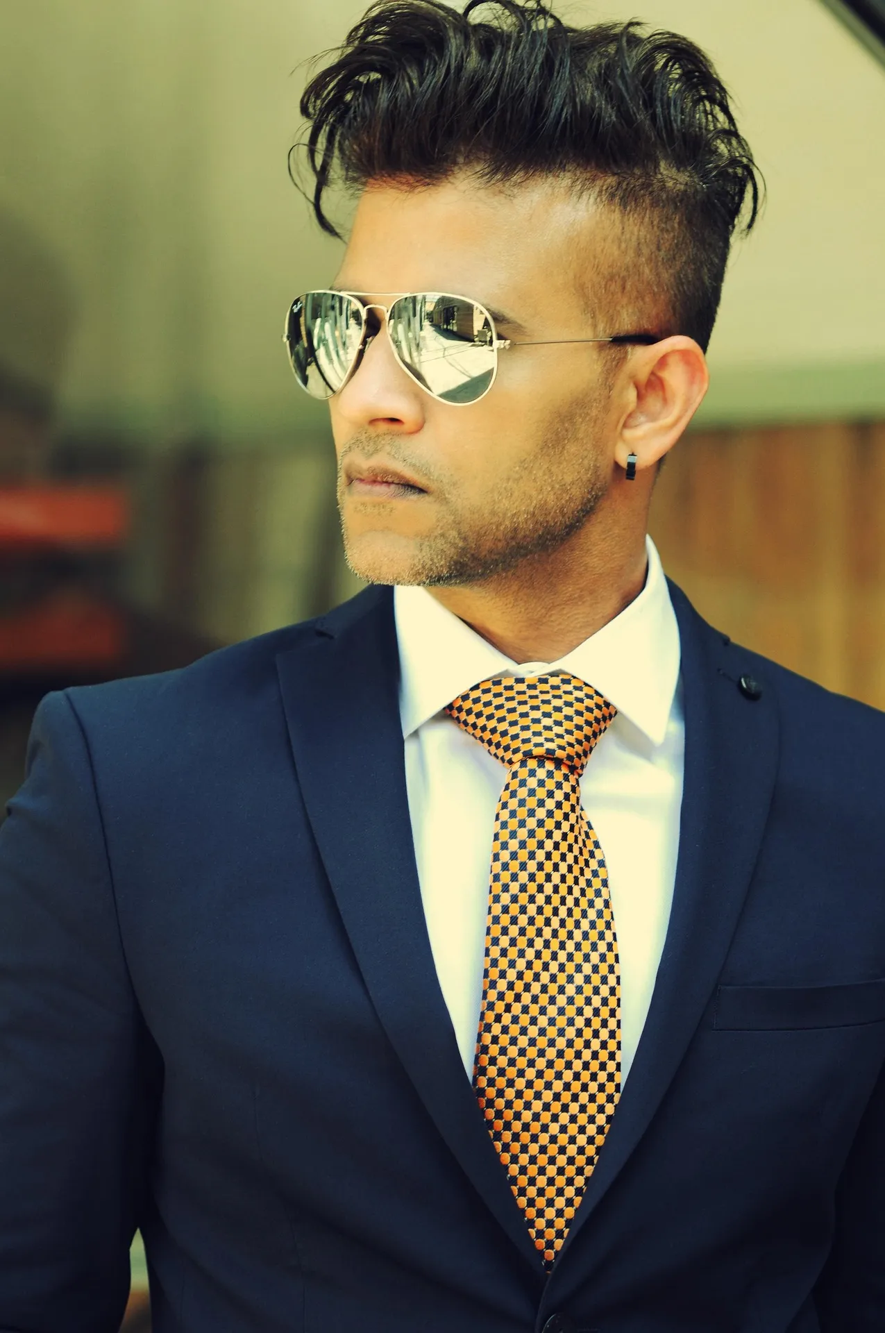 A stylish man in a navy blue suit and gold-checkered tie wearing aviator sunglasses, looking to the side with a confident expression.