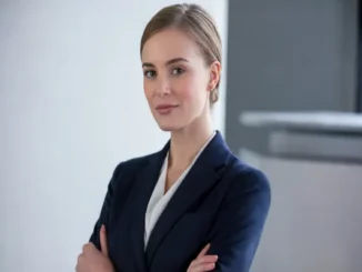A young woman in a sharp navy blue blazer and white blouse standing with arms crossed, looking at the camera with a confident and approachable expression in an office setting.