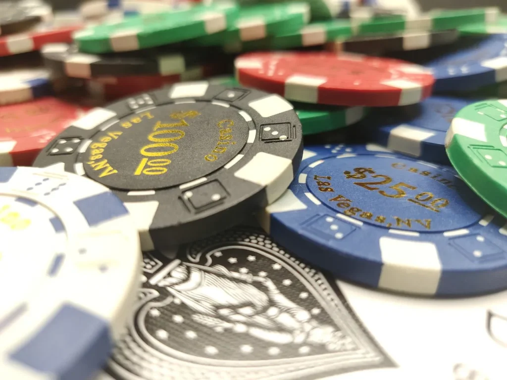 A close-up, top-down view of various colored poker chips, including black $100 chips and blue $25 chips, with "Las Vegas, NV" inscribed on them, along with a portion of a playing card with a spade design, all piled together.