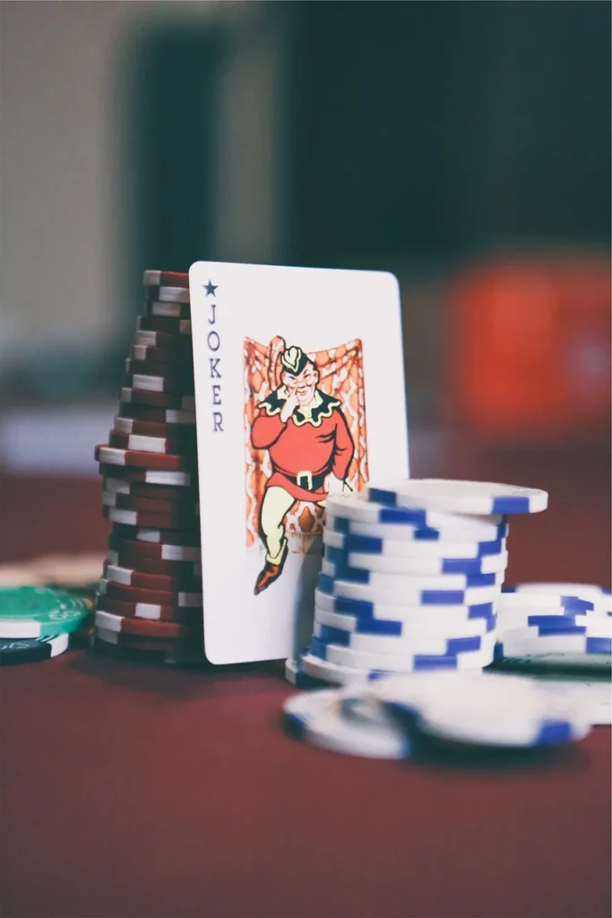 A close-up shot of a Joker playing card standing upright on a red poker table, flanked by tall stacks of red and white poker chips and other scattered chips, suggesting a high-stakes game.