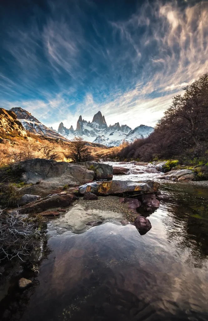A breathtaking wide-angle view of the iconic Mount Fitz Roy and its surrounding snow-capped peaks in Patagonia, Argentina, reflected in a clear, rocky stream with dramatic cloud formations in the sky.