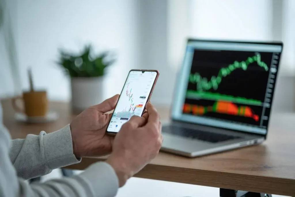 A person holds a smartphone displaying a financial chart while a laptop in the background shows a live trading graph, symbolizing online investing and digital trading courses.