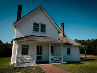 White house with a porch and brick walkway, surrounded by greenery, under a clear blue sky.