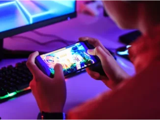 Hands holding a smartphone displaying a video game, with a gaming monitor and RGB keyboard visible in the background.