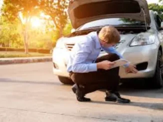 A man in a light blue shirt and dark pants is crouching and examining the front damage of a silver car with its hood open, while holding a tablet.