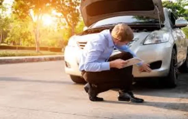 Inspecting Car Damage After Accident A man in a light blue shirt and dark pants is crouching and examining the front damage of a silver car with its hood open, while holding a tablet.