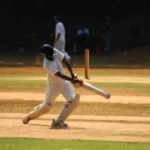 A cricketer in white uniform and helmet plays a shot on a dry, earthy pitch, with a wooden bat.