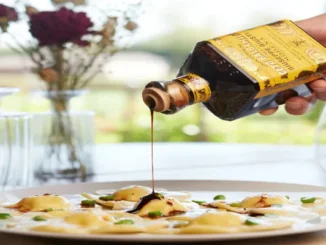 Hand pouring dark, viscous balsamic vinegar from a labeled bottle onto a plate of ravioli.