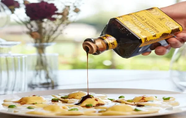 Drizzling High-Quality Balsamic Vinegar Hand pouring dark, viscous balsamic vinegar from a labeled bottle onto a plate of ravioli.