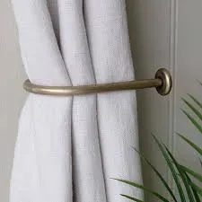 A close-up of a light-colored, possibly linen, curtain held back by a sleek, brushed brass or gold-toned metal U-shaped tieback mounted on a light-colored wall. A hint of green foliage is visible on the right.