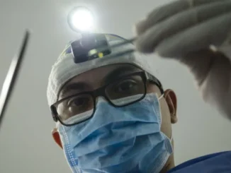 A close-up, low-angle shot of a male healthcare professional (dentist or surgeon) wearing a surgical mask, glasses, and a headlamp, holding medical tools, looking down at the viewer.