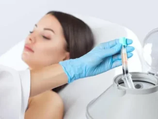 A person in blue gloves holding a test tube with separated blood plasma, next to a machine, with a patient's face blurred in the background.