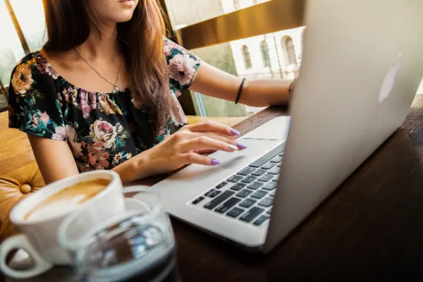 A woman in a floral top works on a silver laptop, with a cup of coffee beside her.