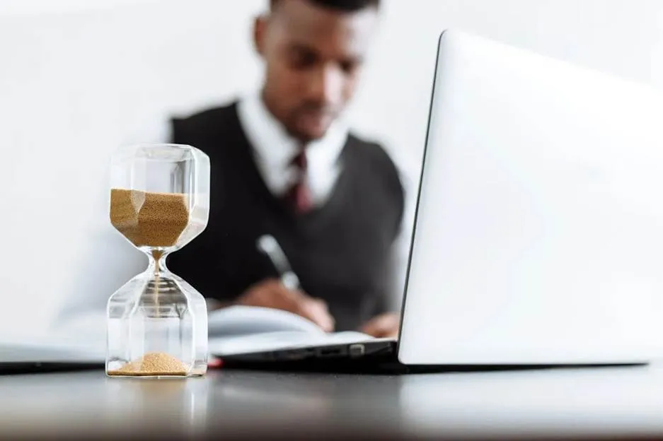 Sand timer in the foreground with a blurred businessman using a computer and taking notes on paperwork, suggesting the importance of timely tax solutions.