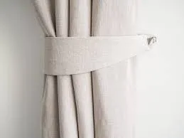 A close-up of a light beige or off-white linen curtain held back by a matching fabric tieback that wraps around the curtain and is secured with a visible silver-toned clip or fastener.