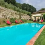 A vibrant blue swimming pool surrounded by a green lawn, red lounge chairs, and a traditional Tuscan villa with a stone wall and trees behind it.