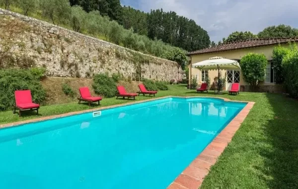 Luxury Villa with Pool in Tuscany A vibrant blue swimming pool surrounded by a green lawn, red lounge chairs, and a traditional Tuscan villa with a stone wall and trees behind it.