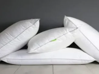 A group of five fluffy white pillows with grey piping, some stacked and some leaning, against a dark grey wall and light wooden floor, representing comfortable bedding options.