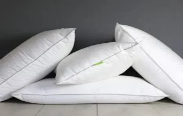 Assortment of Premium White Pillows A group of five fluffy white pillows with grey piping, some stacked and some leaning, against a dark grey wall and light wooden floor, representing comfortable bedding options.