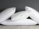 A group of five fluffy white pillows with grey piping, some stacked and some leaning, against a dark grey wall and light wooden floor, representing comfortable bedding options.