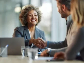 A smiling businesswoman with curly hair shaking hands with a client across a conference table, with a laptop and documents visible.