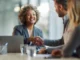 A smiling businesswoman with curly hair shaking hands with a client across a conference table, with a laptop and documents visible.
