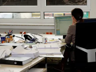 A woman from behind sitting at a desk with numerous stacks of documents and papers, working on a computer.