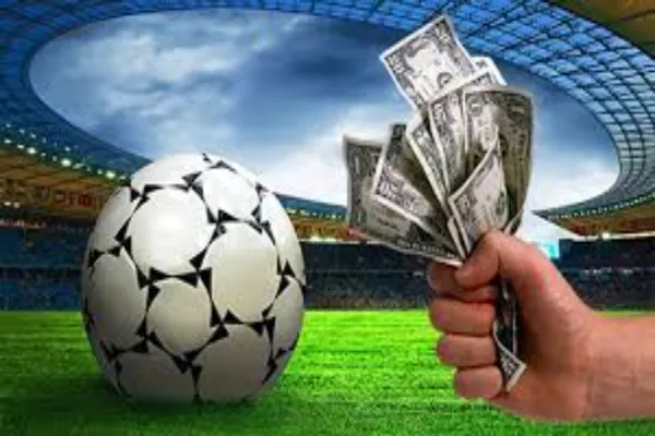 A hand holding a fan of dollar bills next to a soccer ball on a green field inside a stadium.