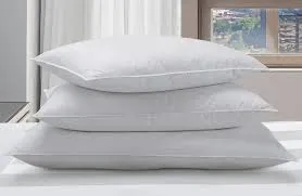 Three white, rectangular pillows are stacked neatly on top of each other, showcasing their loft and clean appearance, with a blurred window and light-colored curtains in the background.