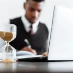 An hourglass with sand running, in sharp focus, with a blurred image of a person in business attire working on a laptop and writing at a desk in the background.