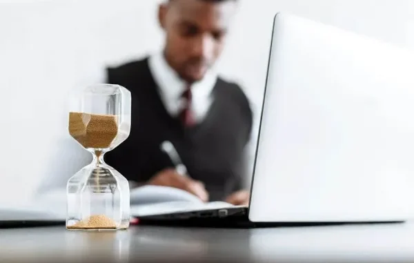Hourglass and Professional Working - Tax Problem Solutions An hourglass with sand running, in sharp focus, with a blurred image of a person in business attire working on a laptop and writing at a desk in the background.