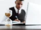 An hourglass with sand running, in sharp focus, with a blurred image of a person in business attire working on a laptop and writing at a desk in the background.