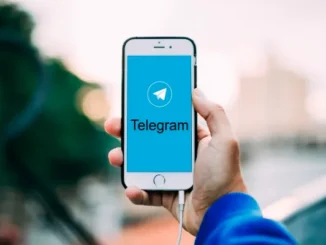 A hand holds a smartphone displaying the Telegram app logo and name on a blue screen, with a blurred outdoor background.