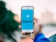 A hand holds a smartphone displaying the Telegram app logo and name on a blue screen, with a blurred outdoor background.