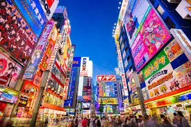 Vibrant night view of Akihabara, Tokyo, with towering buildings covered in brightly lit anime and electronics advertisements, and blurred crowds of people walking below.