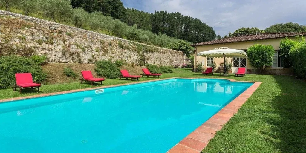 Lush green lawn surrounds a sparkling blue swimming pool at a Tuscan villa, with red lounge chairs inviting relaxation.