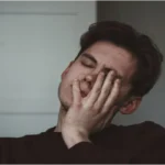 A young man with his eyes closed, rubbing his face with his hand, looking tired or overwhelmed.