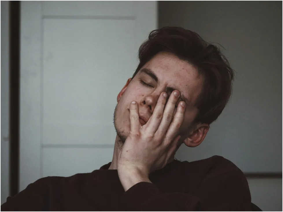A young man with his eyes closed, rubbing his face with his hand, appearing tired or overwhelmed.