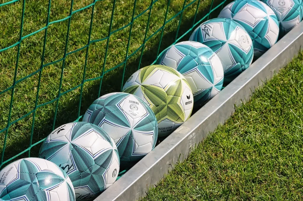 A row of spare soccer balls, mostly white and teal with one yellow, lined up next to a goal net on the edge of a bright green football pitch, representing the tools of the game central to football betting.
