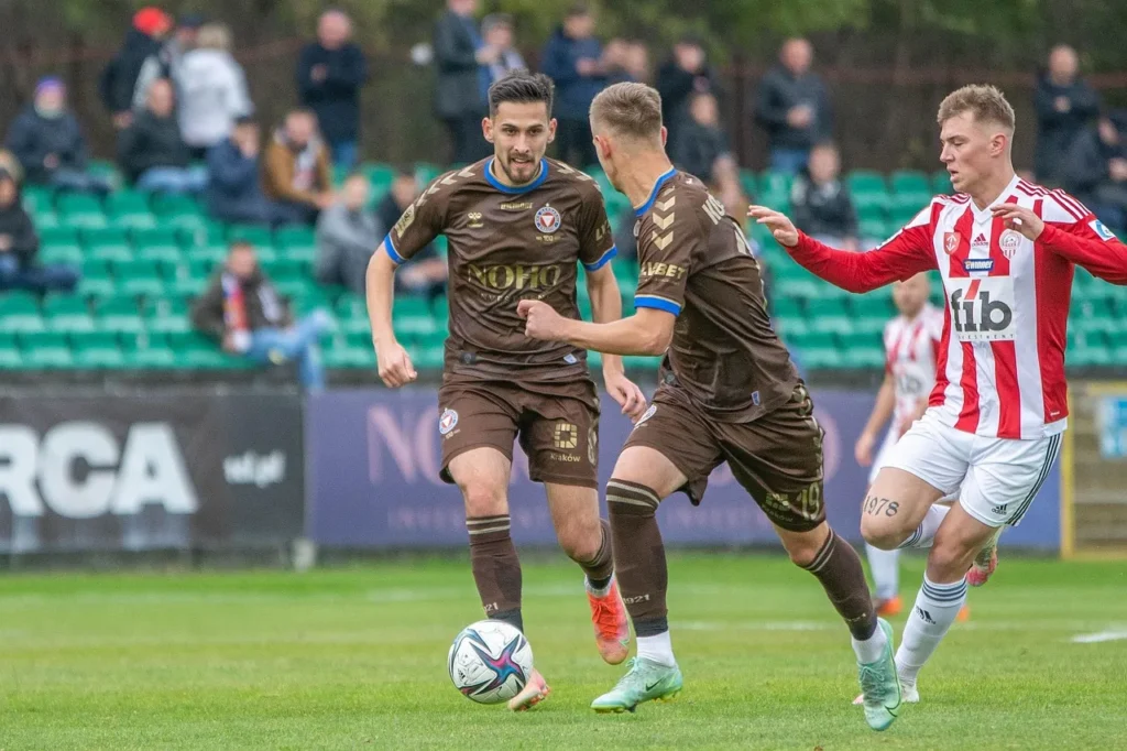 Three football players in different team kits, two in brown and one in red and white stripes, actively competing for the ball on a green pitch during a live match, with spectators in the background, illustrating in-play football action.