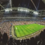 A wide-angle view from behind a crowd of spectators in a massive, brightly lit football stadium during a night match, with the green pitch and players visible, symbolizing the live action crucial for in-play betting.