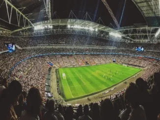 A wide-angle view from behind a crowd of spectators in a massive, brightly lit football stadium during a night match, with the green pitch and players visible, symbolizing the live action crucial for in-play betting.