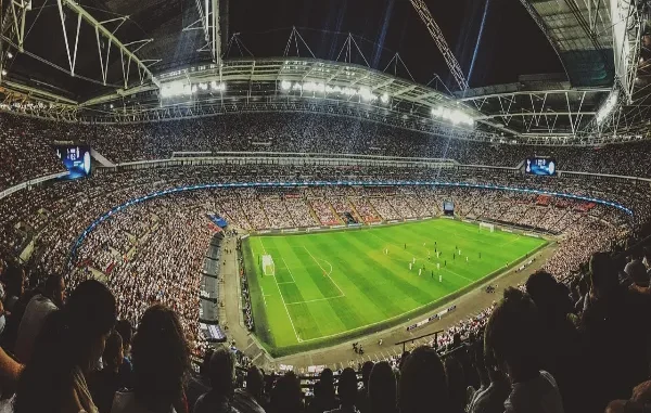 Live Football Betting: How to Make Money When the Match is Already Underway A wide-angle view from behind a crowd of spectators in a massive, brightly lit football stadium during a night match, with the green pitch and players visible, symbolizing the live action crucial for in-play betting.