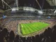 A wide-angle view from behind a crowd of spectators in a massive, brightly lit football stadium during a night match, with the green pitch and players visible, symbolizing the live action crucial for in-play betting.