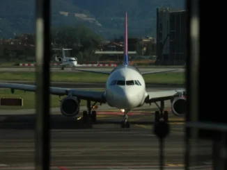 A passenger airplane on the runway at an airport, seen through a window, with a view of mountains and another smaller plane in the background.