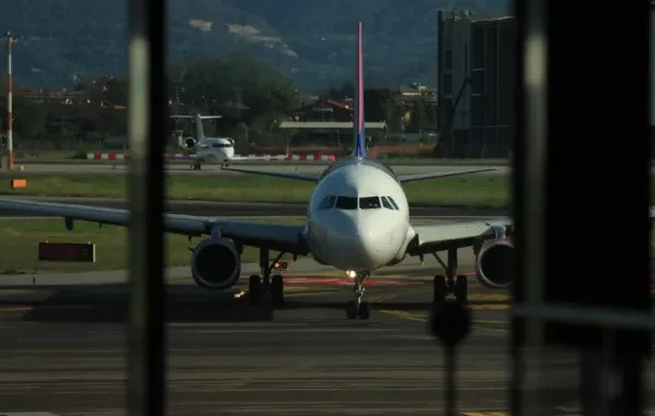 A passenger airplane on the runway at an airport, seen through a window, with a view of mountains and another smaller plane in the background.