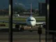 A passenger airplane on the runway at an airport, seen through a window, with a view of mountains and another smaller plane in the background.