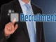 A man in a suit and tie points to a glowing, transparent screen displaying the word "Recruitment," symbolizing digital hiring processes.