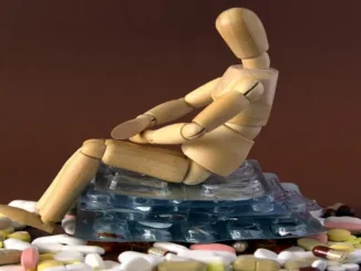 A wooden mannequin figure sits slumped on a pile of various pills and blister packs, symbolizing drug addiction and its burden.