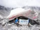 Two people pose with a 'Nepal High Trek Everest Base Camp (5364m)' banner in front of a large, rock marked with red paint at Everest Base Camp.